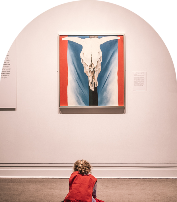 An arch-shaped photograph of a girl wearing a red-vest, seen from behind, kneels on a taupe carpeted floor and looks down presumably at a drawing she is making; she kneels in front of a Georgia O'Keeffe painting against a white wall. The painting depicts a cow’s symmetrical skull floating against what appears to be a cloth backdrop. The backdrop consists of one black vertical band running down the center behind the skull, a blue-white gradient emanating from the center and there is a red vertical band on either edge of the painting.